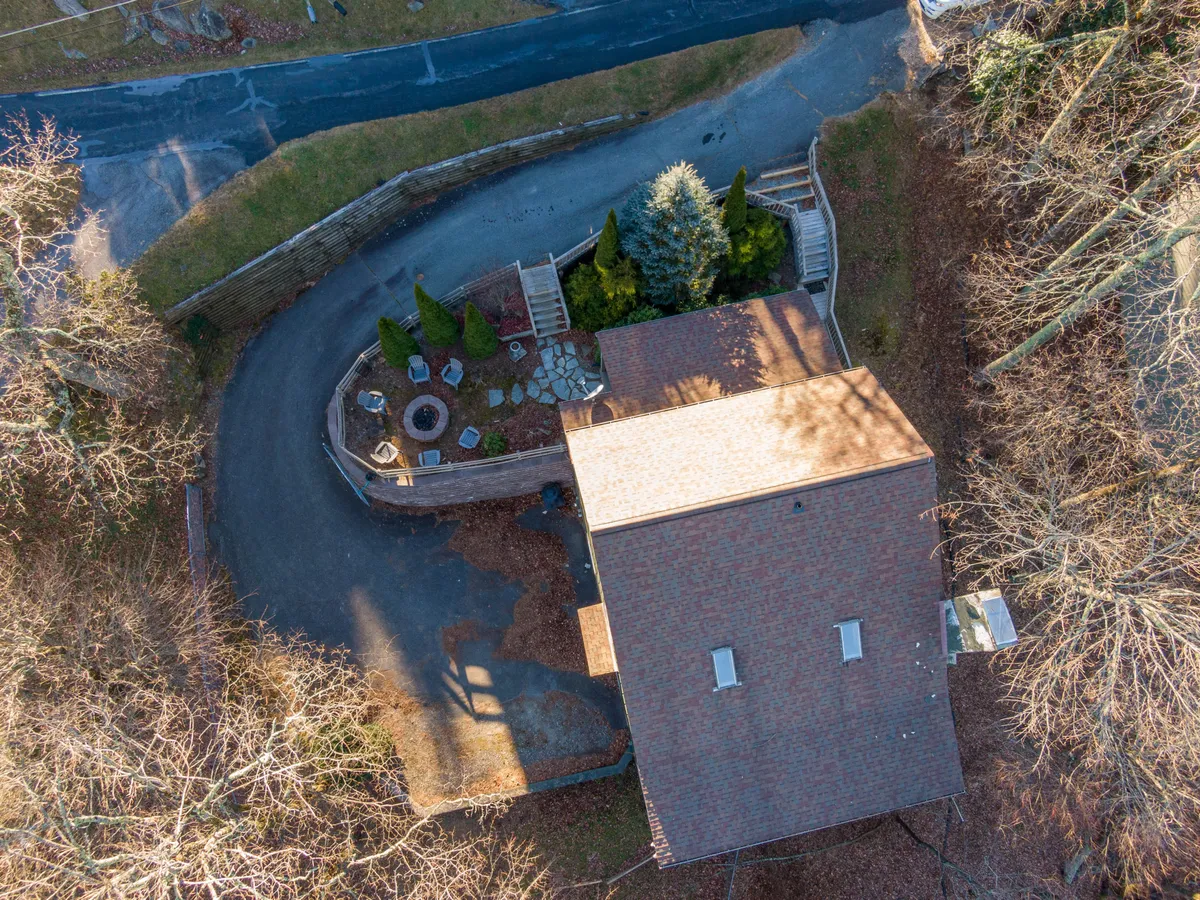 An overhead view of the lodge's roof, a circular stone patio, and a winding driveway, all set amidst dense, mature trees. The spacious outdoor areas are clearly visible, emphasizing the property's connection to nature and its suitability for gatherings or outdoor play.