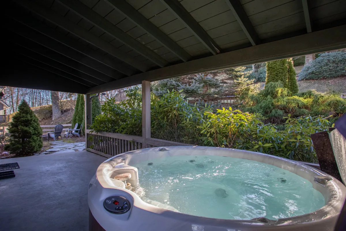 This exterior image showcases a tranquil hot tub surrounded by lush greenery, with comfortable patio seating nearby. The deck space offers an inviting spot to relax, whether enjoying the hot tub, sipping morning coffee, or gathering with friends for summer barbecues. The scene is peaceful and focuses on outdoor enjoyment, with a natural mountain backdrop that changes with the seasons.