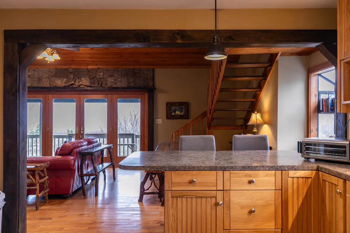 A spacious, open-plan kitchen and seating area is shown, with a red leather sofa, kitchen island, wooden staircase, and large windows bringing in natural light. The view from the windows hints at the mountain setting, creating a direct connection to the outdoors.