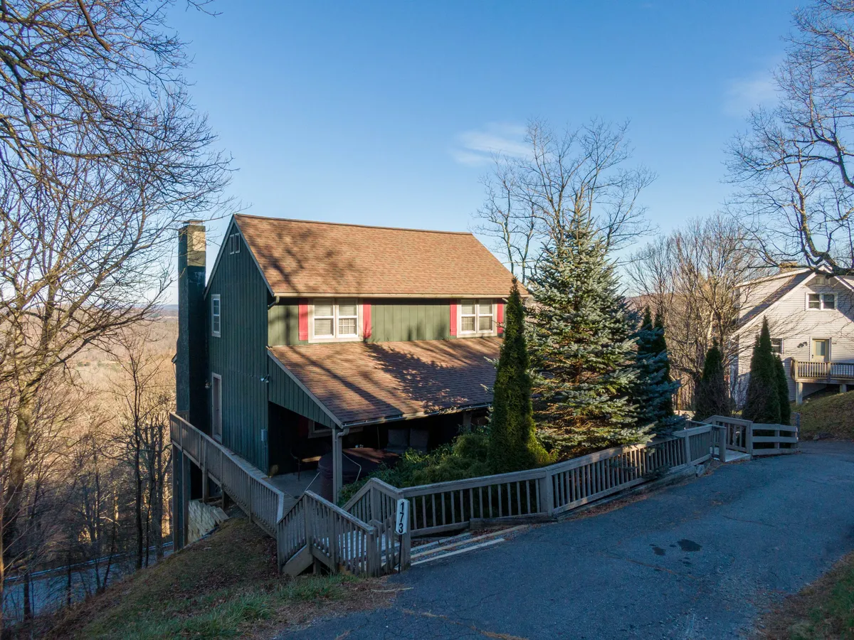 A wide exterior shot of the green house with its brown roof, surrounded by tall evergreens and featuring a wooden railing leading up to the house. The image highlights the expansive, fenced yard ideal for pets, and the inviting porch perfect for watching sunsets and hosting family gatherings.