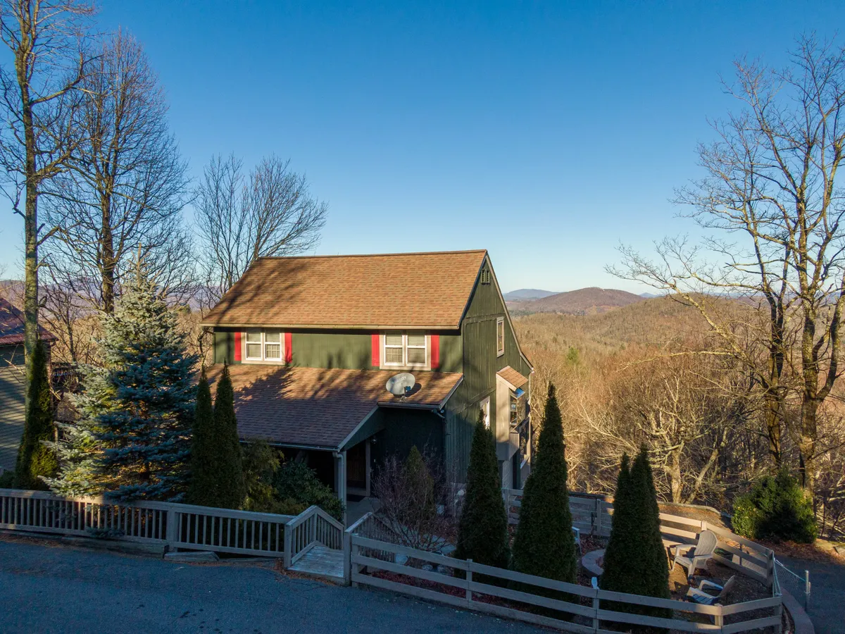 This image features the exterior of Antlers Lodge, showcasing the green house with distinctive red window shutters and a welcoming outdoor seating area nestled among tall conifer trees. The setting feels peaceful and highlights the natural beauty of the surroundings, suggesting opportunities for enjoying the mountain air and seasonal changes.
