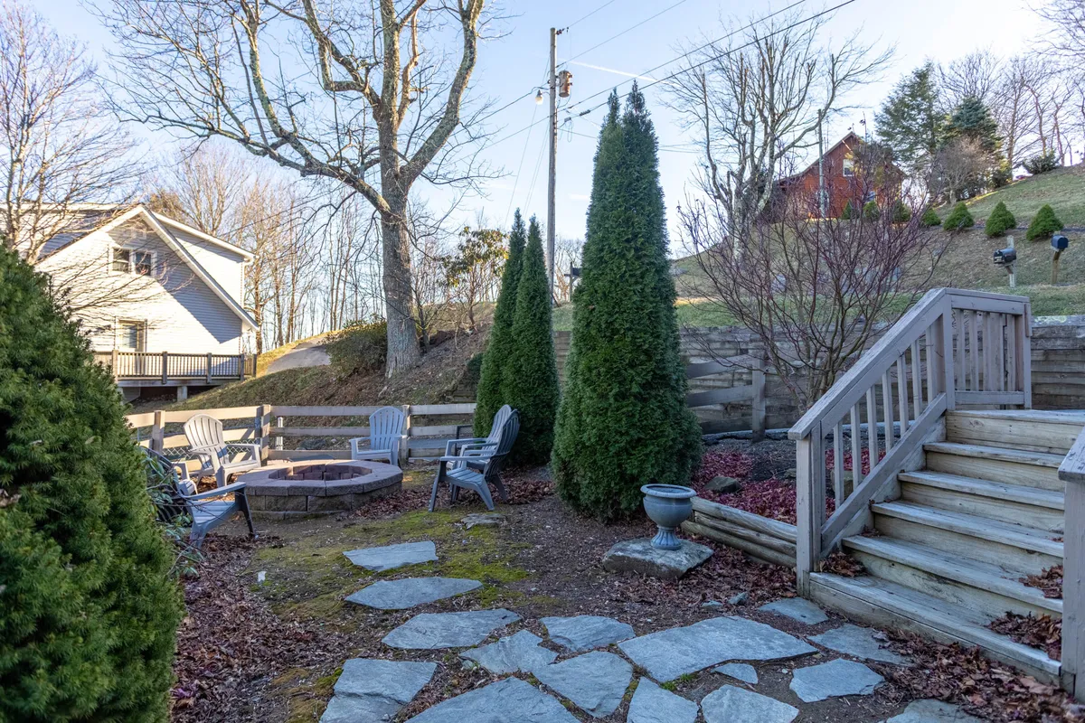 This image displays a cozy outdoor fire pit area surrounded by a stone pathway, ample seating, and encircled by lush evergreen trees. The setup invites groups to gather around the fire, with scenic natural elements that highlight the property's outdoor charm.