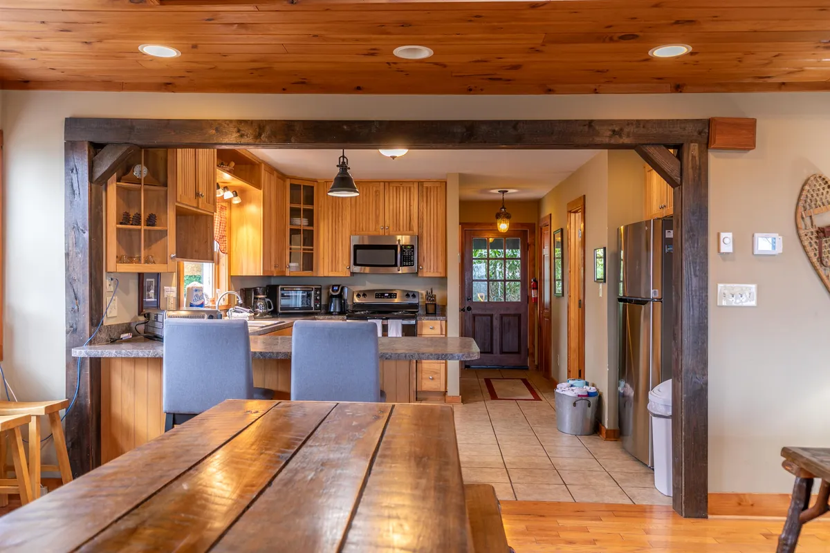 The open dining area features a long wooden table with ample seating, illuminated by natural light from an adjacent doorway that leads out to the covered deck. The kitchen island and part of the living area can be seen in the background, emphasizing the open-concept layout and communal atmosphere.