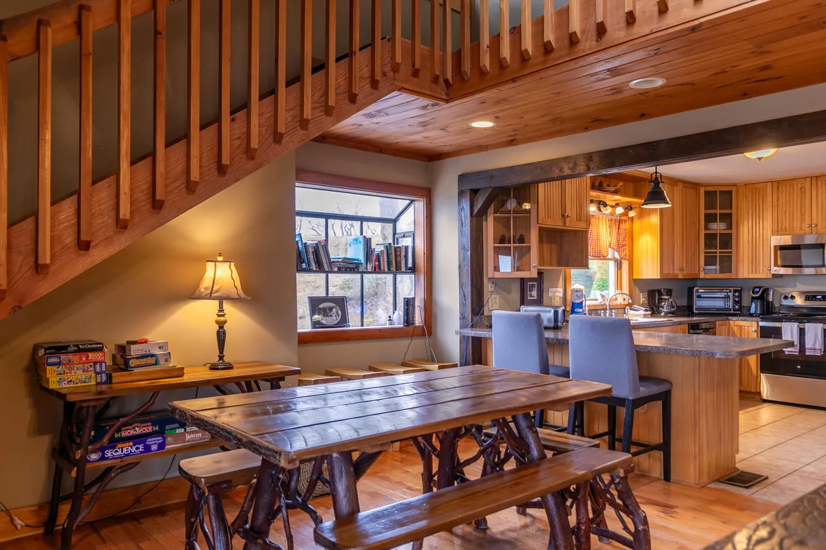 A cozy dining area with a rustic wooden table sits adjacent to the kitchen. A staircase and a window with bookshelves add charm, blending the dining and cooking spaces seamlessly for a warm, collective atmosphere.