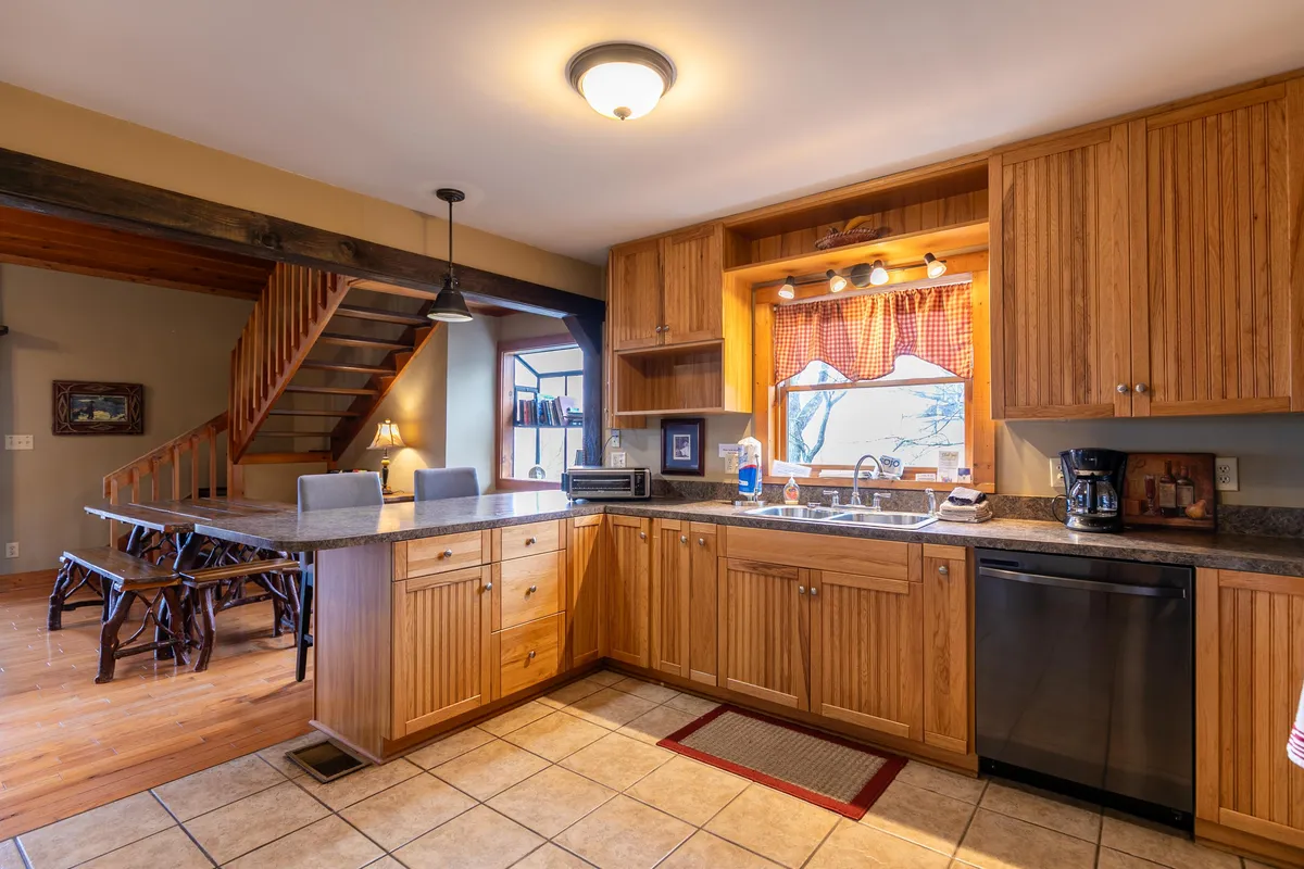This image showcases a well-lit kitchen space featuring stainless steel appliances, wooden cabinets, a kitchen counter, a window adorned with a checkered curtain, and a staircase leading upstairs. The layout highlights both the workspaces and the inviting atmosphere ideal for meal prep and family gatherings.