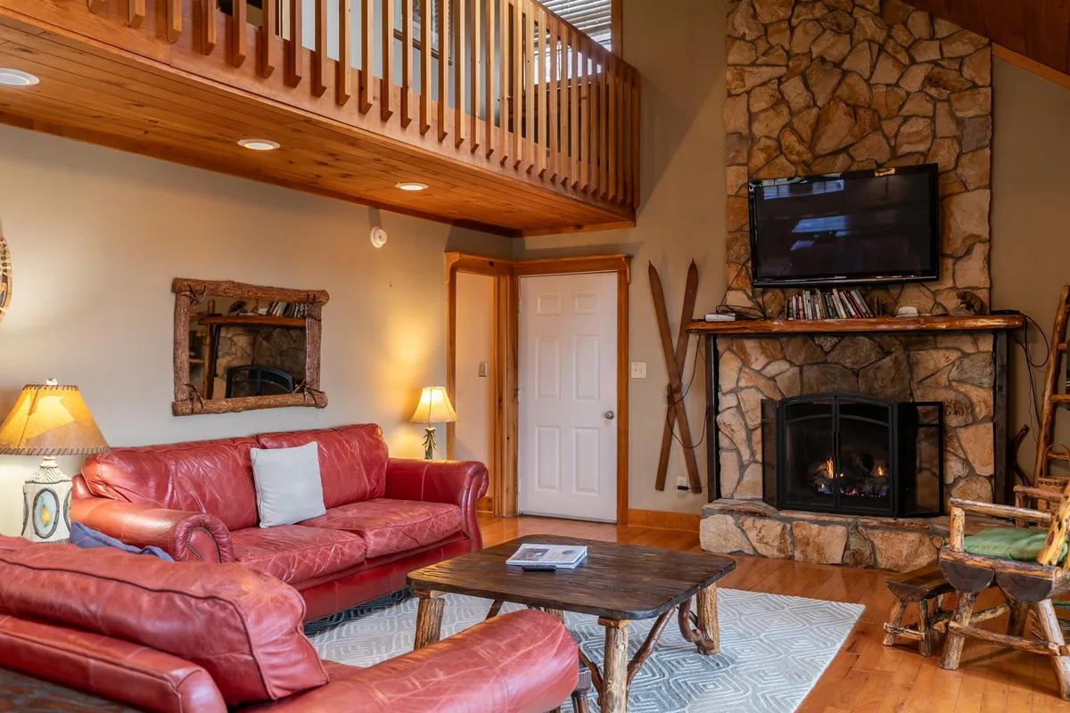 This image features a cozy living area with a red leather sofa facing a stone fireplace, exposed wooden beams above, and a flat-screen TV. The warm, inviting ambiance is accentuated by rustic decor and plush furnishings, suggesting a comfortable place to relax after outdoor adventures.