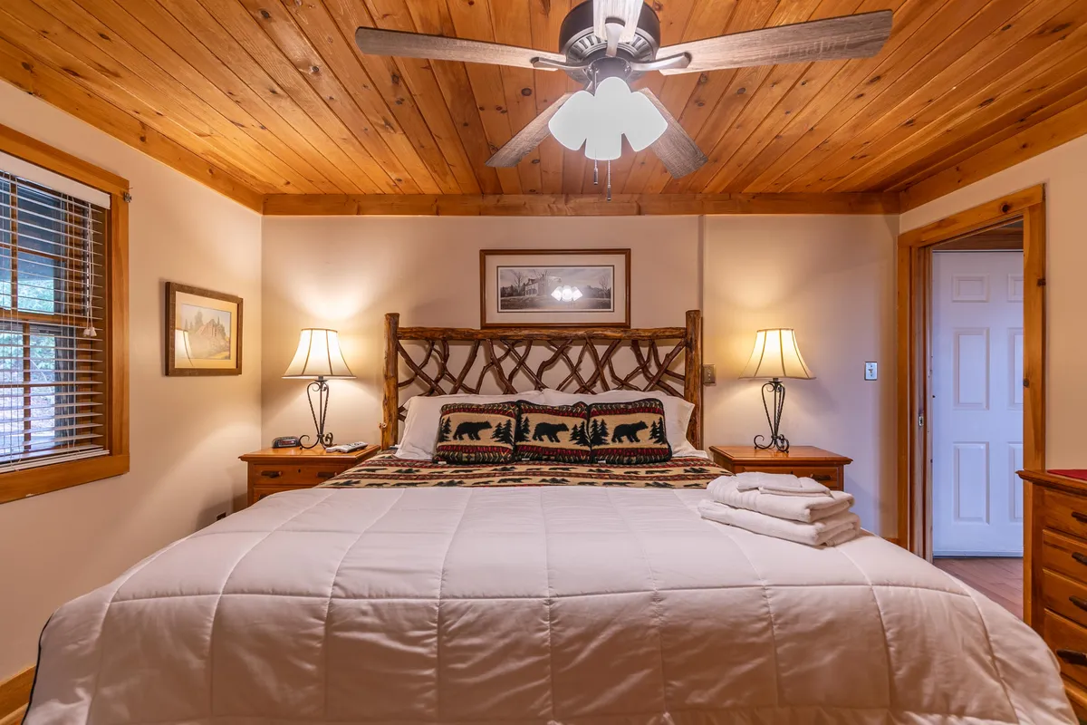 This image features a wooden bed frame with bear-themed pillows, nightstand lamps, and a wooden ceiling, all exuding a warm and welcoming mountain lodge vibe. The rustic decor and cozy linens speak to the idea of retreat and relaxation after a day of adventure.