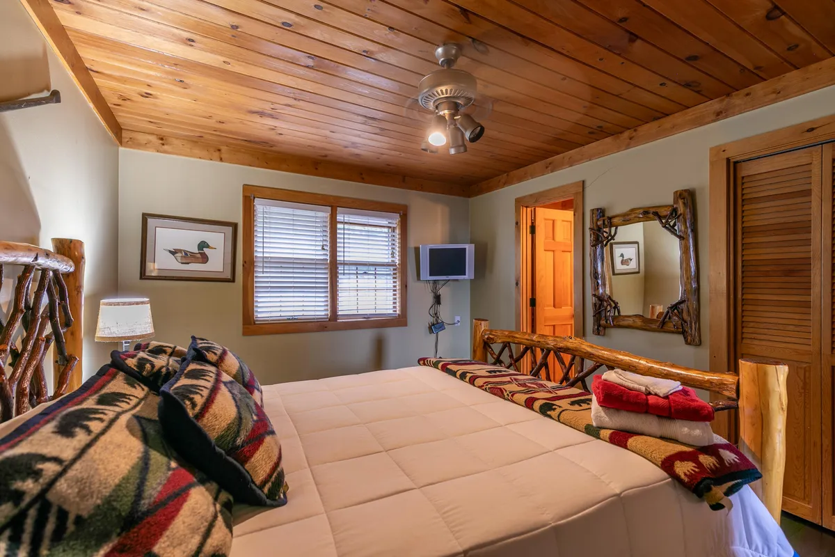 A serene bedroom with a bed adorned with decorative pillows, a wooden ceiling, framed artwork, and a flat-screen TV. The room is filled with soft light, suggesting comfort and peace—perfect for winding down.
