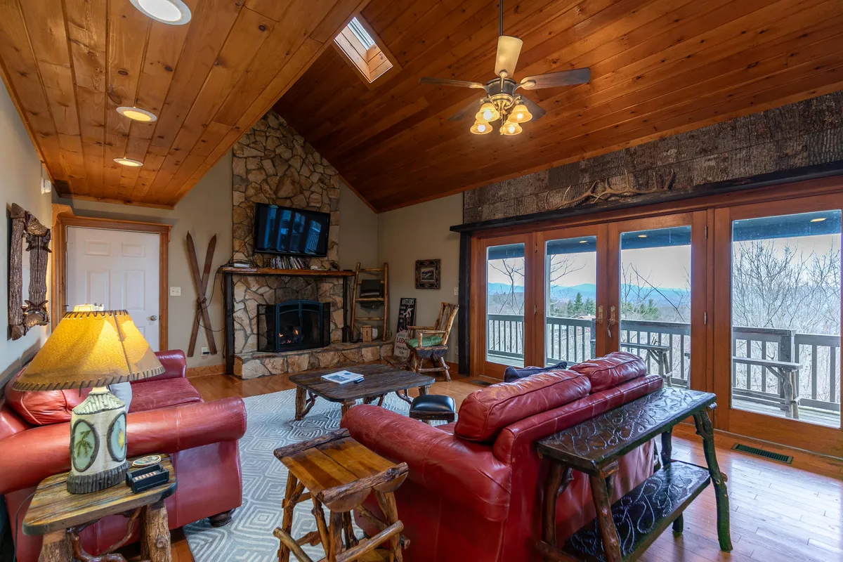 This image centers on the dramatic two-story stone fireplace flanked by large windows offering sweeping mountain views. Plush red leather sofas and a wooden coffee table create a warm, inviting space under soaring timber ceilings, with sunlight streaming in. Decorative lamps add a cozy touch, and part of the deck can be glimpsed through the windows, connecting the interior to the outdoors.